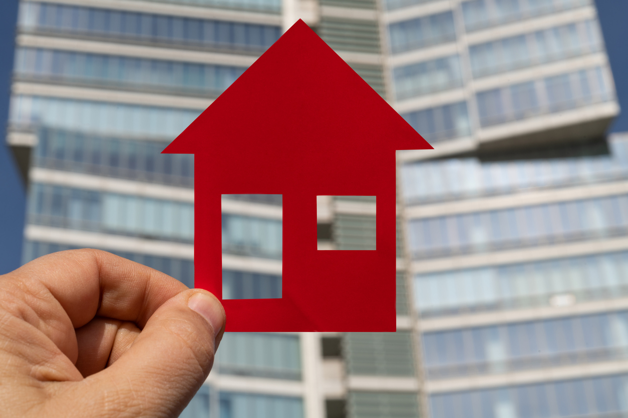 Hand holding a red house icon in front of a modern multi-story building, representing the growth in the Multi-family Industry
